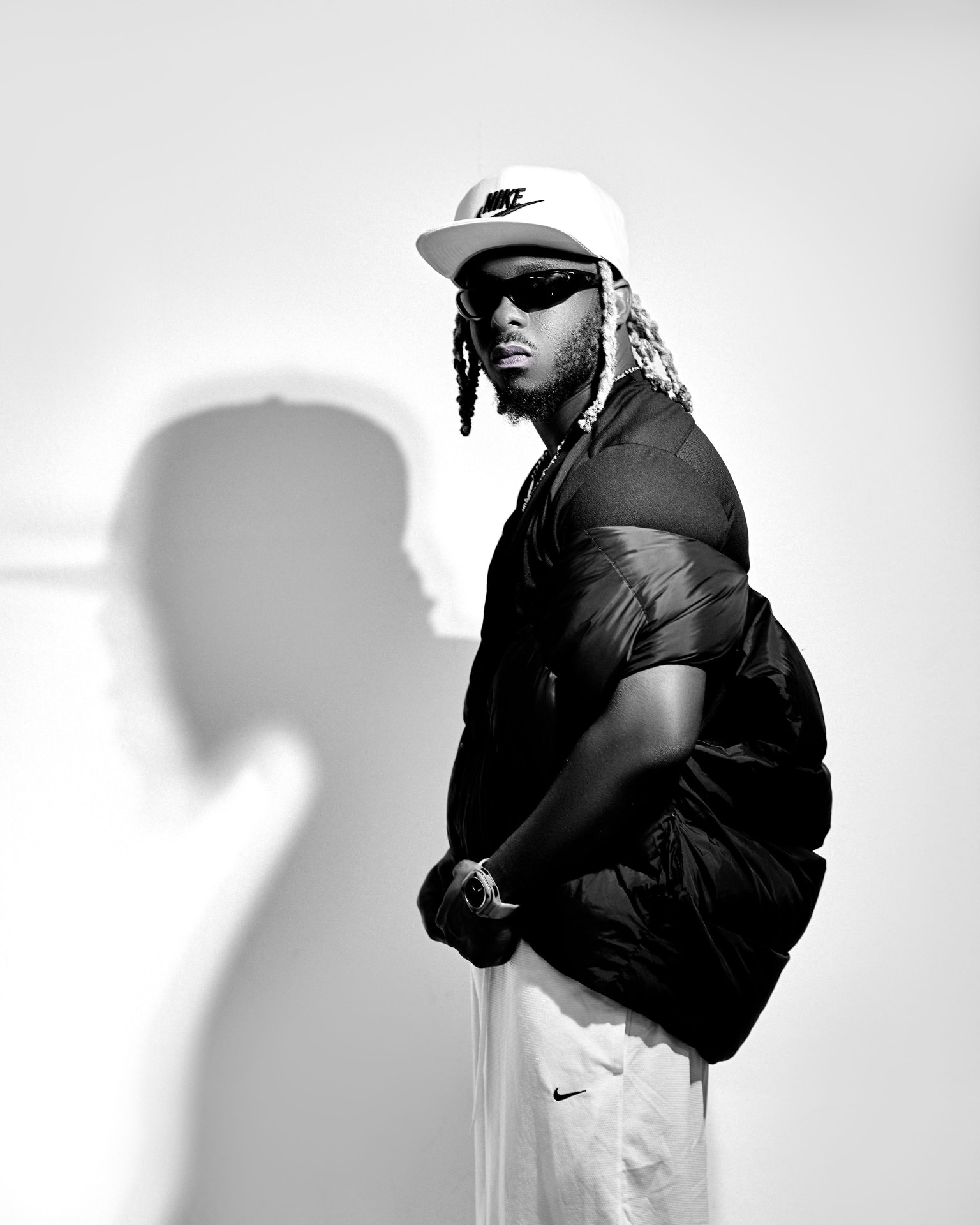 Max Meezy in a black-and-white studio portrait: white Nike cap, dark sunglasses, bleached dreadlocks, a black tee under a quilted black vest and white shorts, standing before a pale backdrop with a soft shadow behind him